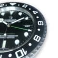 Close-up of a black analog watch face with white hour markers and a black rotating bezel marked with white numerals, showing the time approximately 1:10 with a green arrow-tipped second hand and a date window displaying the 18th, on a white background.