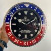 Hand holding a large wall clock designed like a Rolex GMT-Master II watch face with blue and red bezel, showing the time as 10:11:30 and date 23.