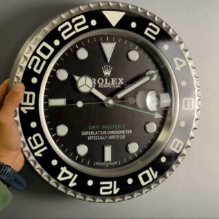 Hand holding a large wall clock designed to resemble a Rolex GMT-Master II wristwatch with black dial, silver markers, and a silver and black bezel featuring 24-hour markings, against a plain grey background.