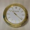 Round wall clock with a gold fluted frame, white face, gold rectangular hour markers, gold hour and minute hands, and the Rolex logo with crown at the top centre.