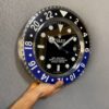 Hand holding a large wall clock designed to resemble a Rolex GMT-Master II watch with a black and blue bezel and black dial against a grey textured wall.