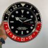 Close-up of a large Rolex GMT-Master II wall clock with a black and red bezel, black dial, white hour markers, and a date window showing 22, held by a person's left hand wearing a watch and a green sleeve against a plain background.