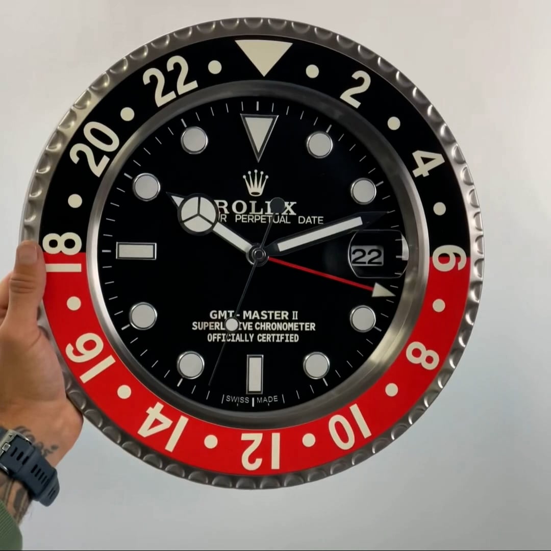 Close-up of a large Rolex GMT-Master II wall clock with a black and red bezel, black dial, white hour markers, and a date window showing 22, held by a person's left hand wearing a watch and a green sleeve against a plain background.