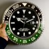 Hand holding a large wall clock designed to look like a Rolex GMT-Master II watch, featuring black and green bezel with white numerals and a black face with white hour markers and hands, showing the time as 10:09:31 and the date as 6.