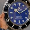 Close-up of a blue Rolex Submariner wall clock with large white hour markers and hands, showing the time at approximately 10:10, being held by a hand on the left side against a grey textured background.