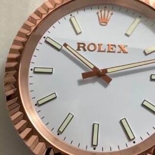 A close-up of a round wall clock with a white face, gold hour markers, hands, and a rose-gold scalloped frame, displaying the time as 10:10 held by a hand wearing a black glove against a textured light background