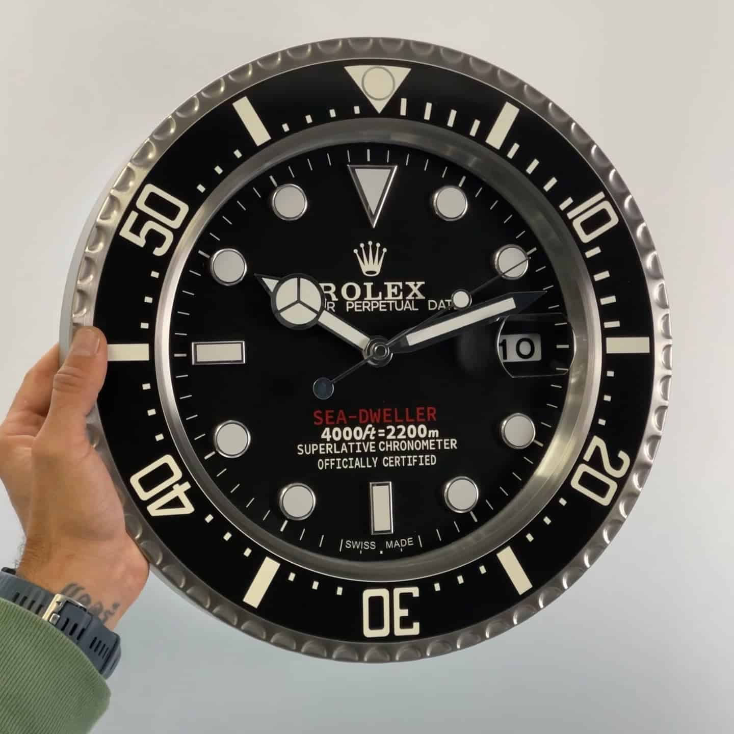 A hand holding a large wall clock designed to look like a Rolex Sea-Dweller dive watch face with black dial, white hour markers, and silver bezel markings, displaying the time 10:12:45 and date 10.