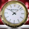 Close-up of a round gold Rolex clock with a white face, black hour and minute hands, and gold hour markers, set against a blurred red and pink background