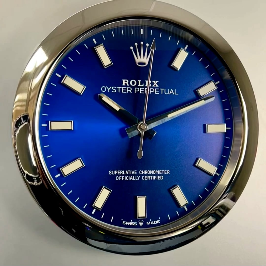 Close-up of a Rolex Oyster Perpetual watch face with a blue dial, silver hour markers, and silver hands showing the time as 10:10:01, with text indicating it is a superlative chronometer officially certified and Swiss made.