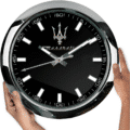 Hands holding a large round clock with a black face, white markers, Maserati logo and branding, and silver frame.