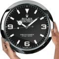 Close-up of a black Rolex Oyster Perpetual Explorer wall clock with white hour markers and hands, held by two hands on either side against a white background