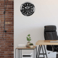 Modern office corner with a black leather chair, wooden desk, small table holding potted plants and stacked books, exposed brick wall, and a black Rolex Explorer wall clock showing 11:10 on a white wall.