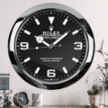 Wall clock designed like a Rolex Oyster Perpetual Explorer watch face with black background and white markers, mounted on a pale wall above a table with a smaller clock, a white mug with a leaf pattern, decorative boxes, and a glass dome with pine cones and dried plants.