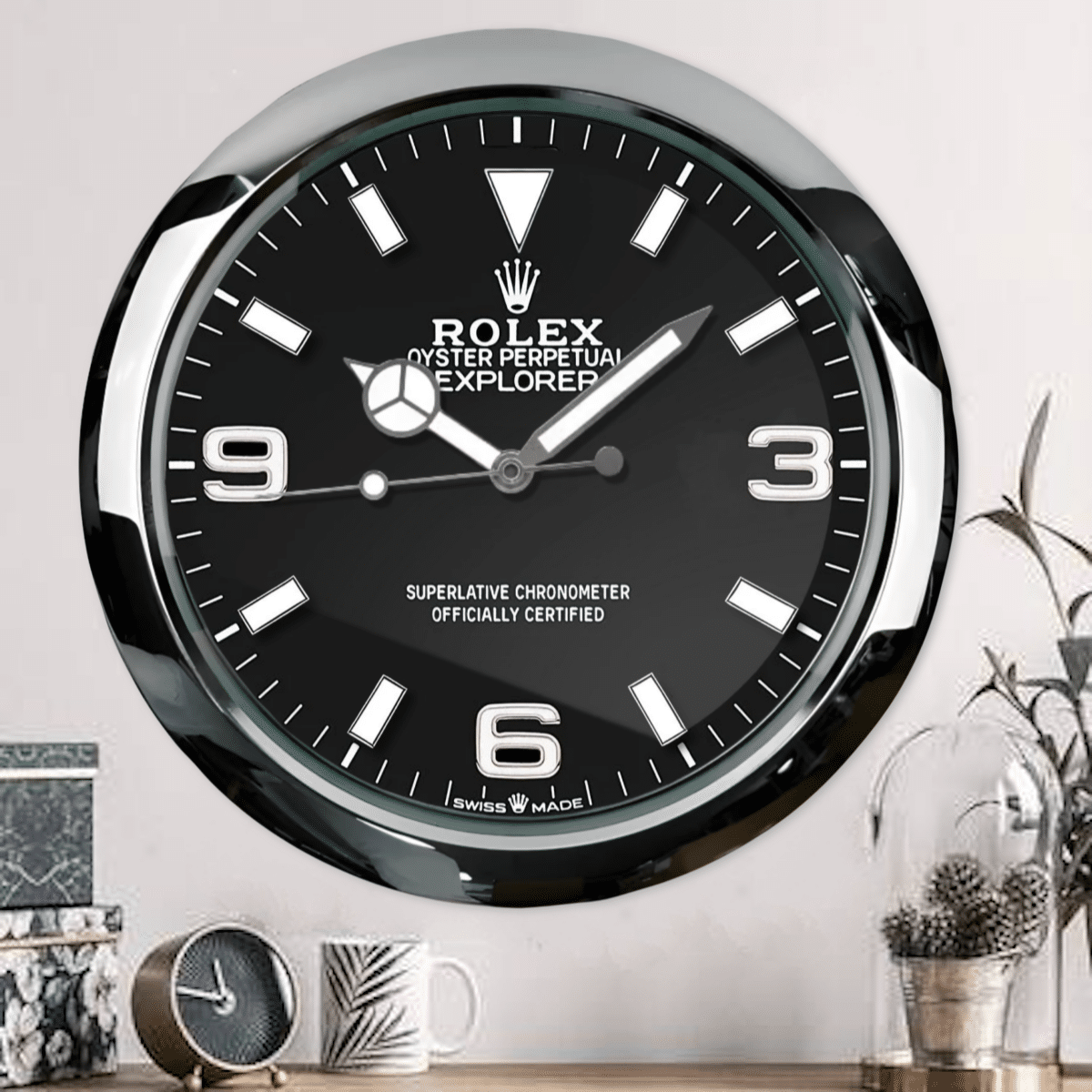 Wall clock designed like a Rolex Oyster Perpetual Explorer watch face with black background and white markers, mounted on a pale wall above a table with a smaller clock, a white mug with a leaf pattern, decorative boxes, and a glass dome with pine cones and dried plants.