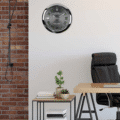 Modern office corner with a black leather chair, wooden desk, small side table holding two potted plants and stacks of books, a wall clock designed like a Rolex watch, and a section of exposed red brick wall with a hanging cord.
