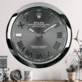 Wall clock designed to resemble a Rolex Oyster Perpetual Datejust watch with Roman numerals and silver detailing, mounted above a wooden shelf holding a small clock, a mug, and decorative items including a glass dome with pine cones and a plant on the right.