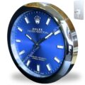 A round wall clock designed to resemble a Rolex Oyster Perpetual watch face, featuring a blue dial with white rectangular hour markers and silver hands, encased in a shiny metallic frame with a modern mounting plate shown separately in the upper right corner.