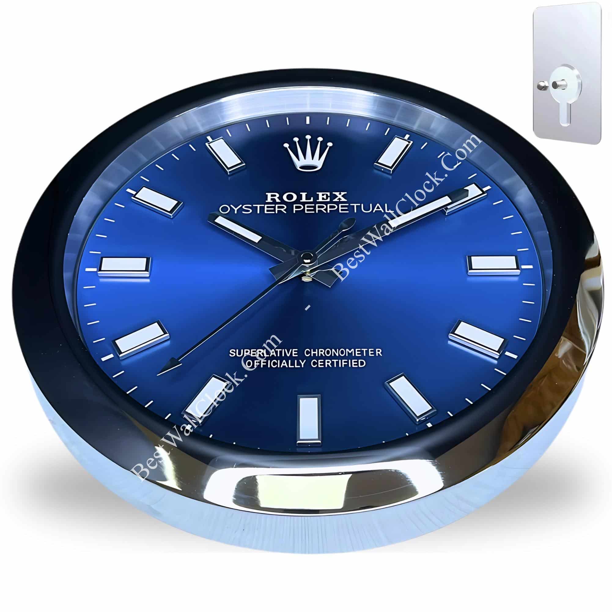 Close-up of a Rolex Oyster Perpetual wall clock with a deep blue dial, silver hour markers, and silver hands, mounted in a shiny chrome frame, showing a modern winding key beside it on a white background.