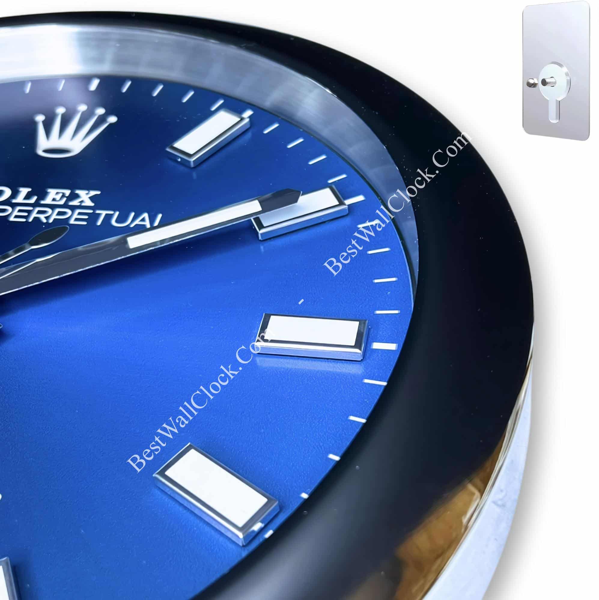 Close-up of a blue Rolex Perpetual wall clock face with rectangular hour markers and silver hands, housed in a black round frame, alongside a silver wall mounting plate with keyhole design on a white background.
