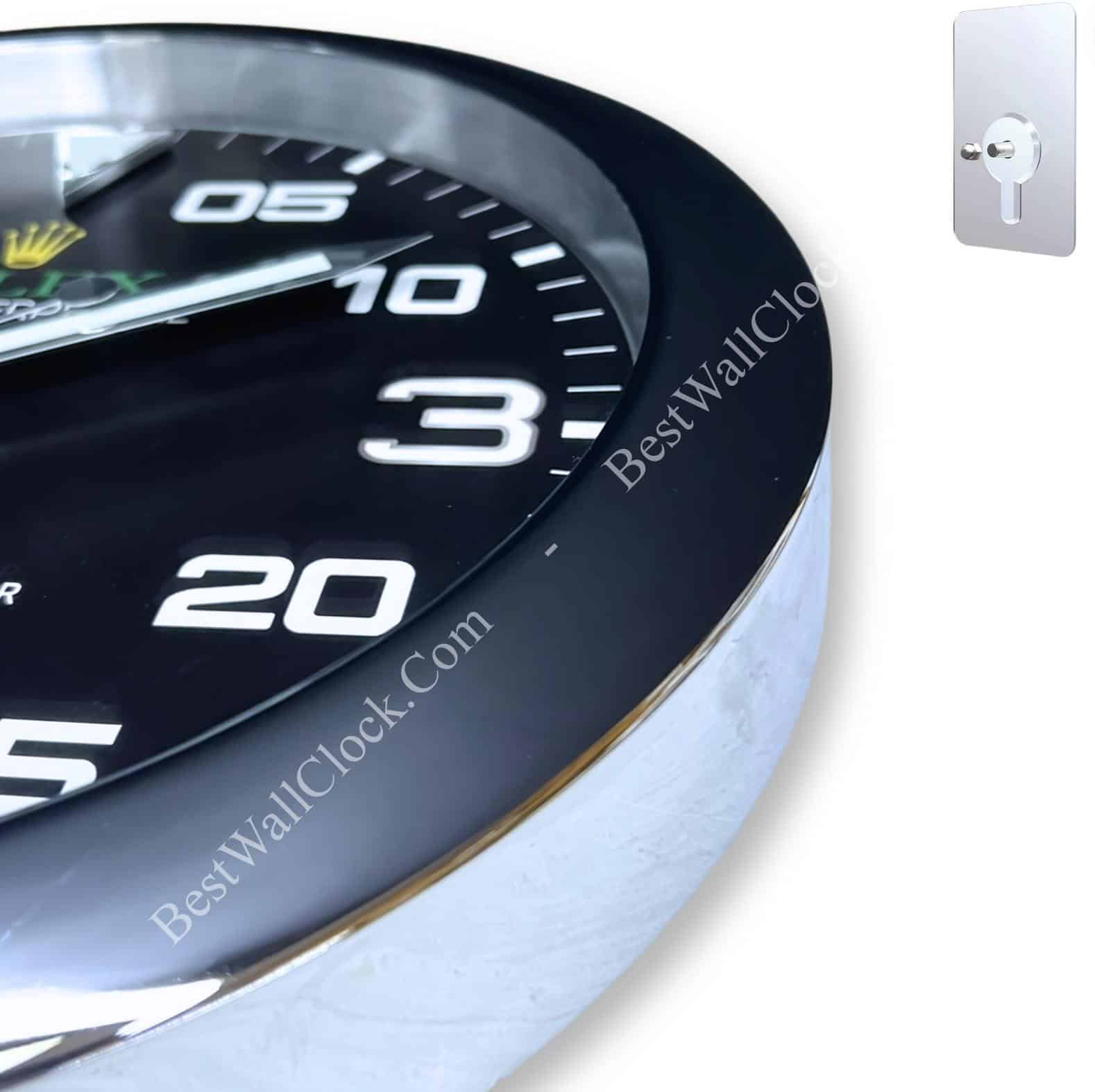 Close-up view of a black wall clock with white numbers and a glossy metallic edge, alongside a small silver wall hook on a white background.