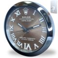 Round wall clock designed like a Rolex watch face with brown background, silver Roman numerals, the Rolex crown logo at the top, and text including 'ROLEX OYSTER PERPETUAL DATEJUST' and 'SUPERLATIVE CHRONOMETER OFFICIALLY CERTIFIED', encased in shiny chrome bezel, shown with a keyhole cover for mounting.