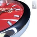 Close-up view of a red Coca-Cola branded wall clock with white hour markers and silver hands, featuring a shiny black and silver frame against a white background.