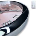 Close-up of a Rolex Oyster Perpetual Datejust wall clock with a pink face, silver Roman numerals, and sleek silver and black clock hands set in a polished black and silver frame, alongside a modern silver wall mounting bracket on a white background.