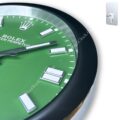 Close-up of a green Rolex Oyster Perpetual watch face with silver and white hour markers and black-and-white hands, featuring the Rolex crown logo near the 12 o'clock position, encased in a round black and silver frame.