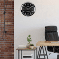 Rolex wall clock with a black face and silver frame hanging on a white wall in a minimalist office setting with a wooden desk, black leather chair, small plants, and stacked books beside a brick wall.