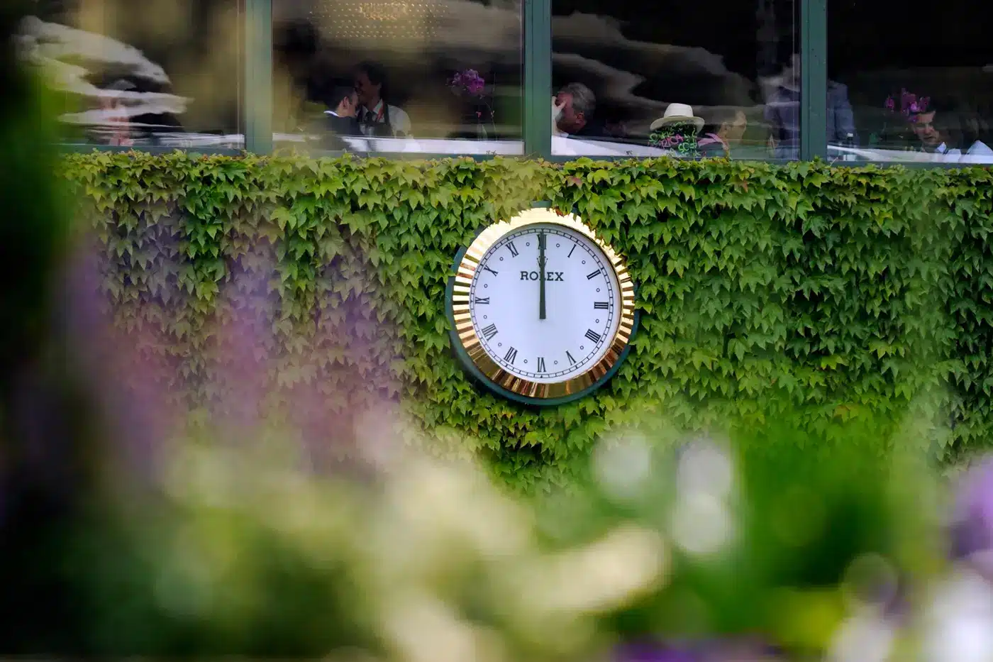 A large Rolex wall clock with a gold rim is mounted on an ivy-covered wall, reading approximately 4:10. Blurred flowers are in the foreground, adding a touch of elegance. Buy Rolex wall clocks at great prices to adorn your space with timeless style.