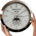Person holding a Patek Philippe wall clock with a silver frame and a white face, featuring day, date, and moon phase indicators, with the phrase
