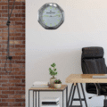 Modern office interior with a black leather chair, a wooden desk, a small table with potted plants and books, a brick wall on the left, and a rolex wall clock with a metallic frame and lime green hands and markers on a white wall.