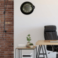 Rolex wall clock designed like a Cartier watch hanging on a white wall above a wooden desk with a black leather office chair, next to a side table with books and potted plants, and a brick wall with a hanging lamp on the left side.