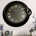 Black rolex wall clock with Roman numerals and blue hands hanging on a white wall above a wooden surface with a small table clock, a patterned mug, decorative boxes, and a glass cloche with dried plants.