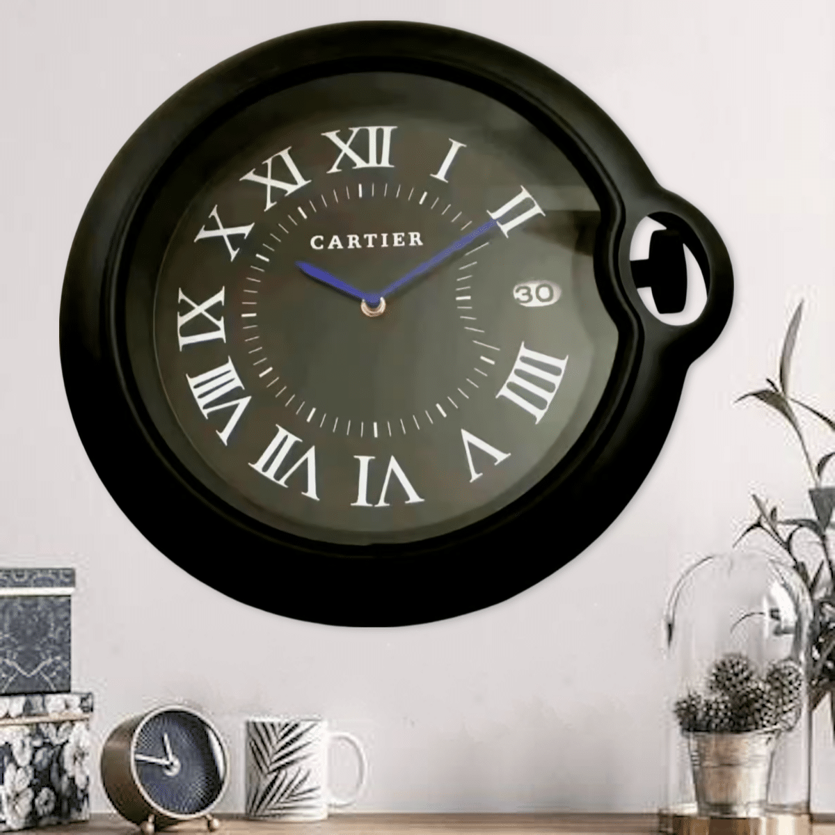 Black rolex wall clock with Roman numerals and blue hands hanging on a white wall above a wooden surface with a small table clock, a patterned mug, decorative boxes, and a glass cloche with dried plants.