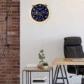 Rolex wall clock with a gold frame and blue dial featuring multiple time indicators, hanging on a white wall next to a workspace with a wooden desk, black office chair, side table with books, and potted plants against a partially brick wall background.