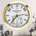rolex well clock featuring a large Patek Philippe wall clock with a gold rim, white face, and calendar functions displayed above a wooden surface with a smaller clock, a mug, and decorative plants