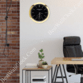 rolex well clock black Patek Philippe wall clock with gold frame hanging above a modern office desk with books and plants against a white and brick wall