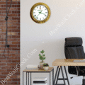 rolex well clock gold-framed wall clock with white face and roman numerals hanging above a wooden desk and black office chair in a modern workspace with brick and white walls