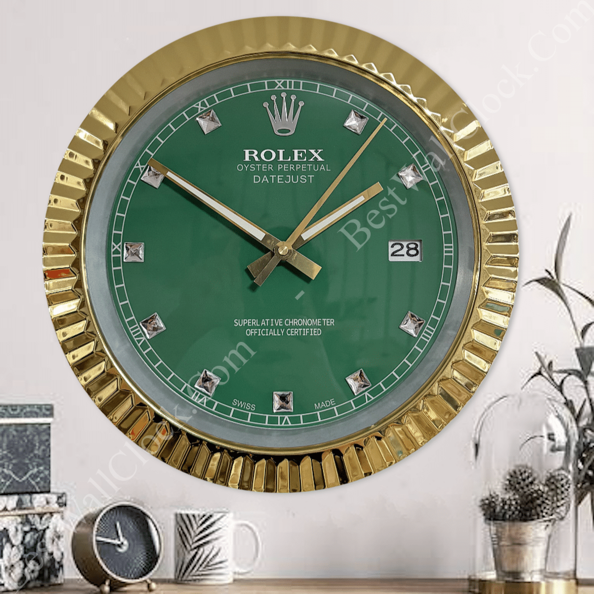 Rolex well clock featuring a green face with gold bezel and diamond hour markers mounted on a wall above a wooden surface with decorative items including a small clock, a patterned mug, and a glass dome with a cactus plant.