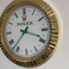 Wall clock with a white face, black Roman numerals, green hands, and a gold bezel, branded with the Rolex logo and crown.