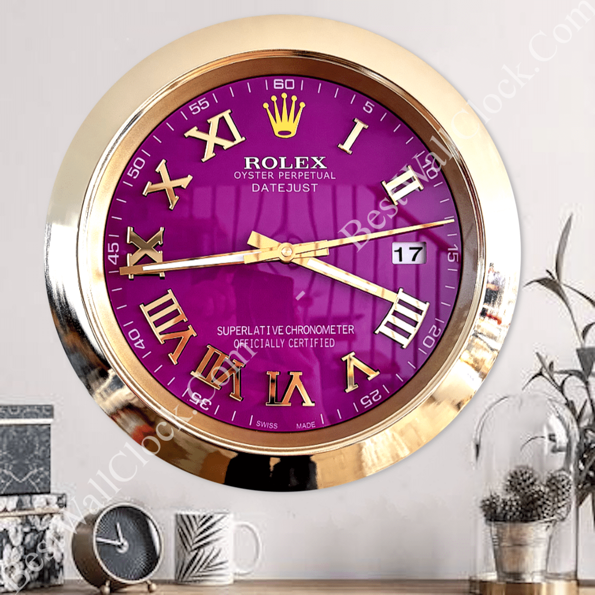 Gold-framed Rolex wall clock with a purple face, gold Roman numerals, gold hands, and a date display showing 17, mounted above a wooden surface with decorative objects including a small clock, a mug, and a glass dome with plants.