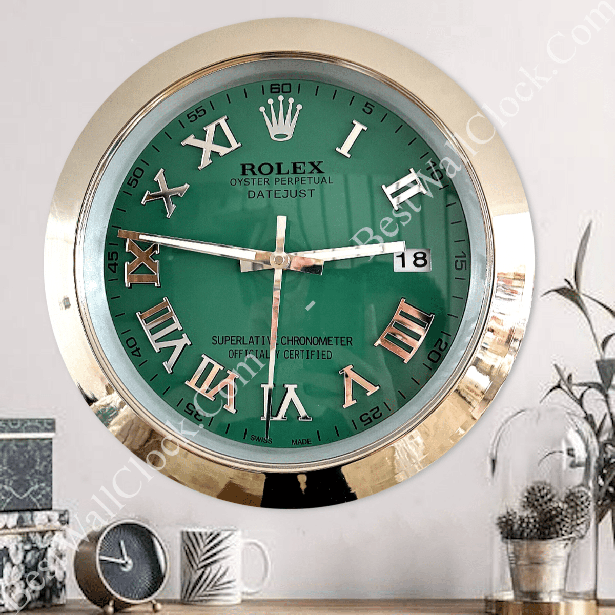 Wall-mounted Rolex-style clock with a green face, gold rim, Roman numerals, and white clock hands, showing the time as 2:47:30, with the date displayed as 18, set above a wooden surface with decorative items including a small black clock, a white mug with a leaf pattern, and a glass dome containing pine cones and a plant.