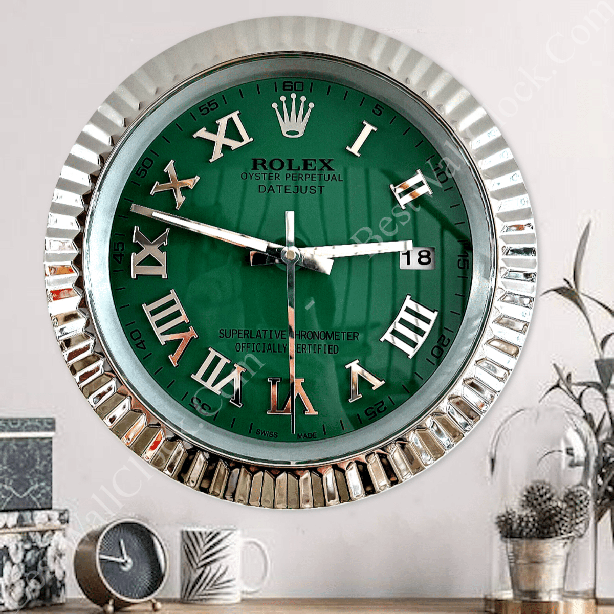 Wall clock designed like a Rolex Oyster Perpetual Datejust watch with a green face, silver Roman numerals, silver bezel, and date display showing the number 18, mounted on a white wall above a wooden surface holding a small clock, a plant pot, and a white mug with a leaf design.