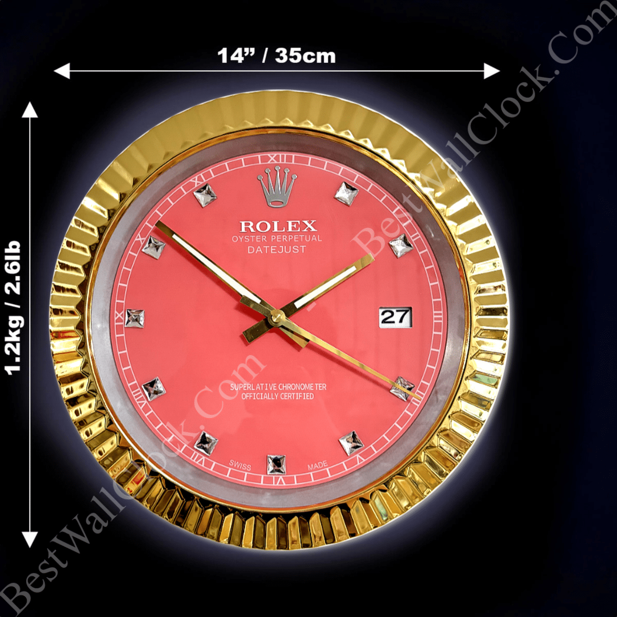 Round wall clock styled like a Rolex Datejust watch with a gold fluted bezel, pink dial, diamond hour markers, gold hands, and a date window showing 27, measuring 14 inches (35 cm) in diameter and weighing 1.2 kg (2.61 lb) against a dark background.