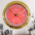 Wall clock designed to resemble a Rolex Datejust watch with a pink face, gold bezel, diamond-shaped hour markers, and gold hands, showing the time as 10:55:20, mounted on a grey wall above a surface with a small clock, a mug, and potted plants.