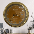 Gold-framed wall clock with a brown face displaying the brand name Rolex, diamond-shaped hour markers, and a date window showing 14, hanging on a light wall above a wooden surface with decorative items including a small round clock, a white mug, and a glass-covered plant pot.
