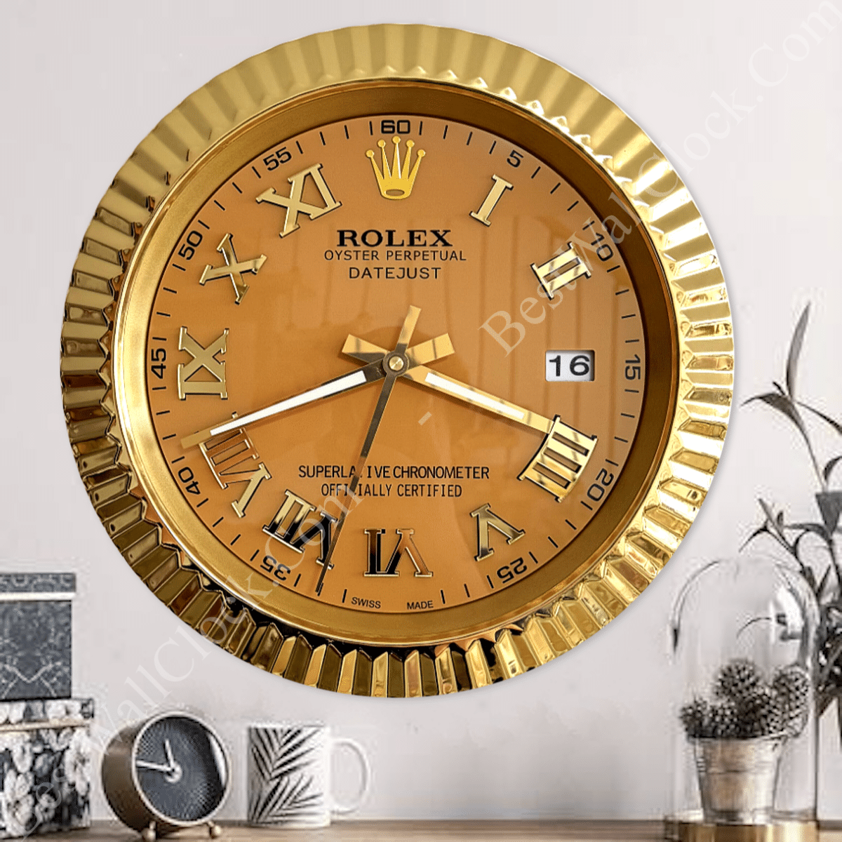 A gold wall clock designed to resemble a Rolex Oyster Perpetual Datejust watch with Roman numeral hour markers, a date window showing the 16th, and gold fluted bezel, mounted on a plain light-coloured wall above a wooden surface holding a small round clock, a mug with a palm leaf design, and a decorative plant under a glass dome.