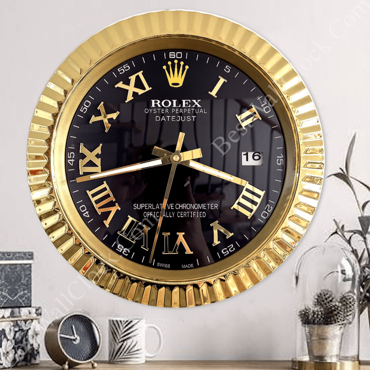 Round wall clock styled like a Rolex watch with a gold fluted bezel, black face, gold Roman numerals, gold hands, and a date window showing 16, mounted on a light wall above a table with a small clock, a patterned mug, and a plant in a glass dome.