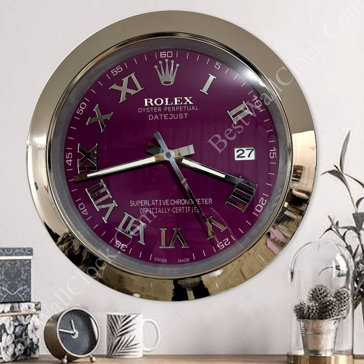 Wall clock with a gold frame and a dark purple face featuring Roman numerals, labeled as Rolex Oyster Perpetual Datejust with a date display showing 27, mounted on a white wall above a wooden surface with small decorative items including a smaller clock, a mug, and a plant in a glass dome.